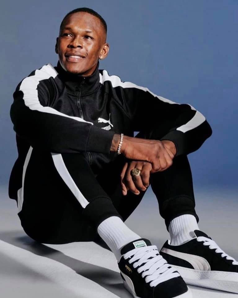 Israel Adesanya in black and white tracksuit against a bluish background