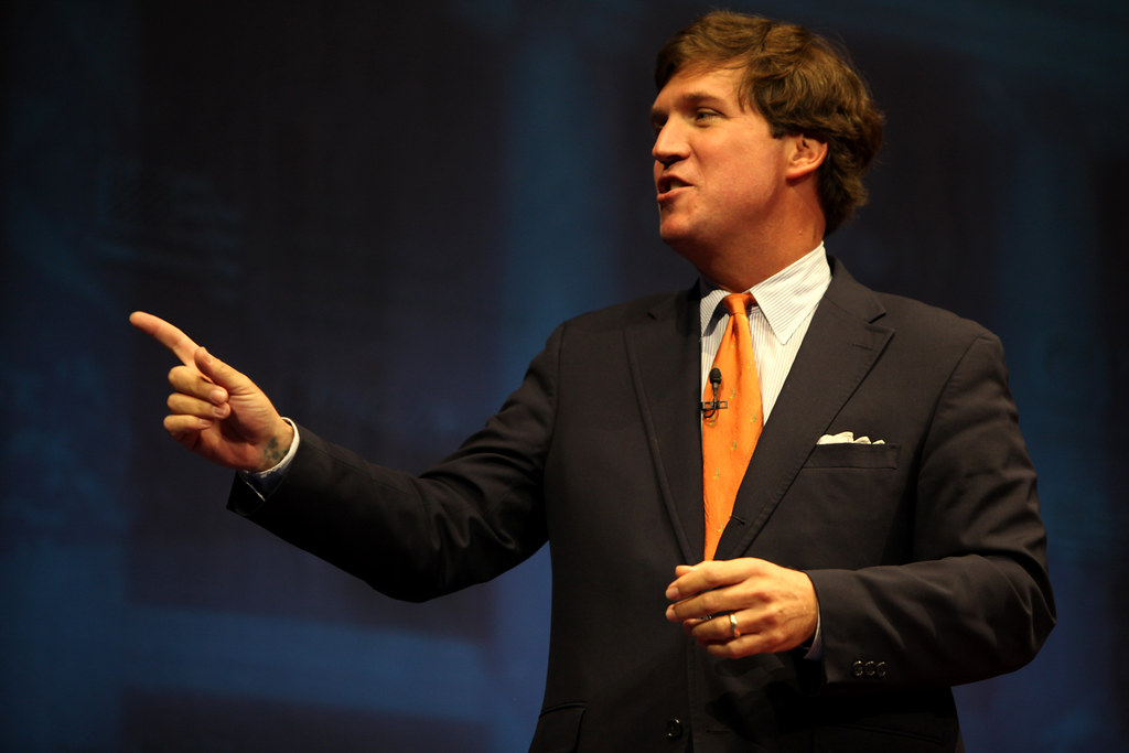 Tucker Carlson in a suit, talking and gesturing on stage