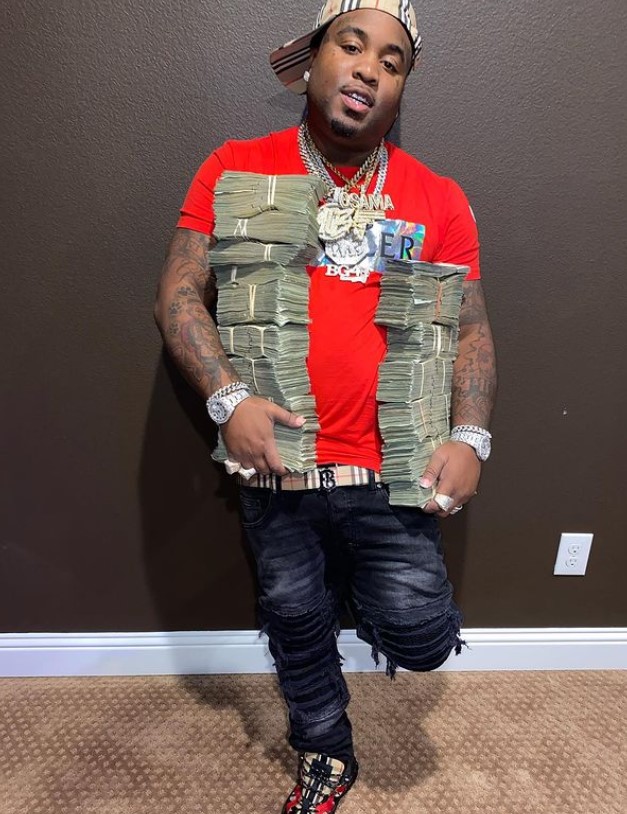 Mo3 standing against a wall holding two stacks of cash, one in each hand