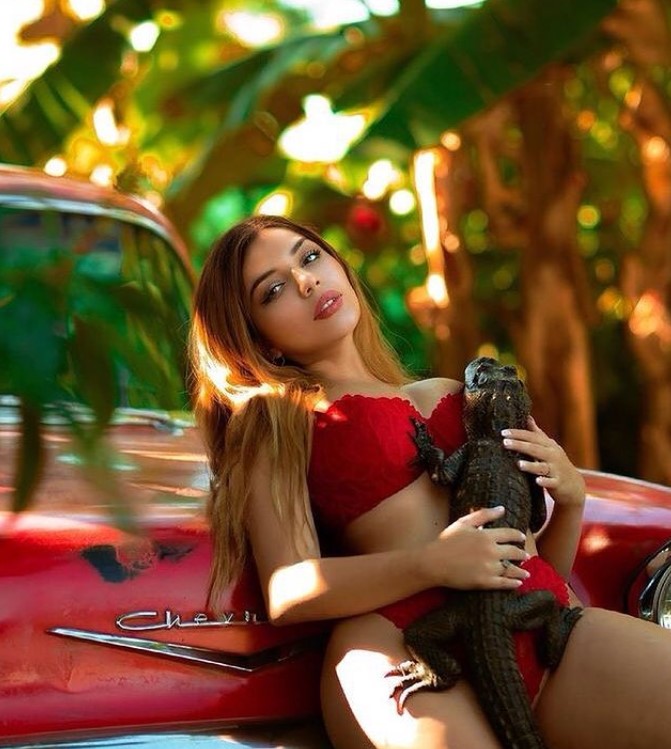 Katiana Kay in red bikini against a red car, holding what looks like a small gator