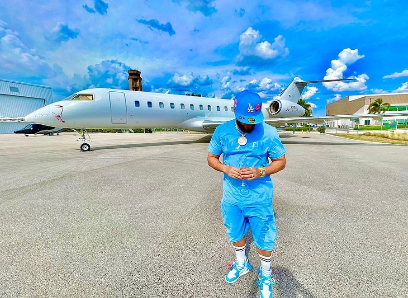 El Alfa looking down, wearing a blue outfit, and standing in front of a plane