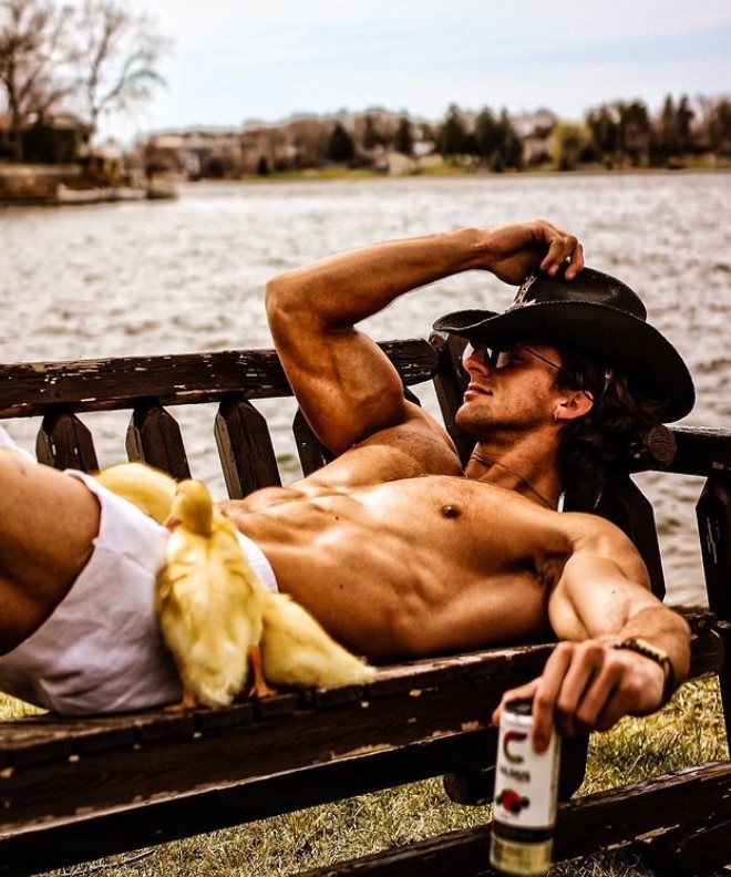 TooTurntTony wearing sunglasses and a cowboy hat, laying on a bench by the water