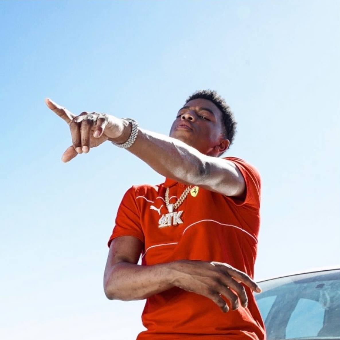 Yungeen Ace in red, positioned with his arm out and pointing, next to a car